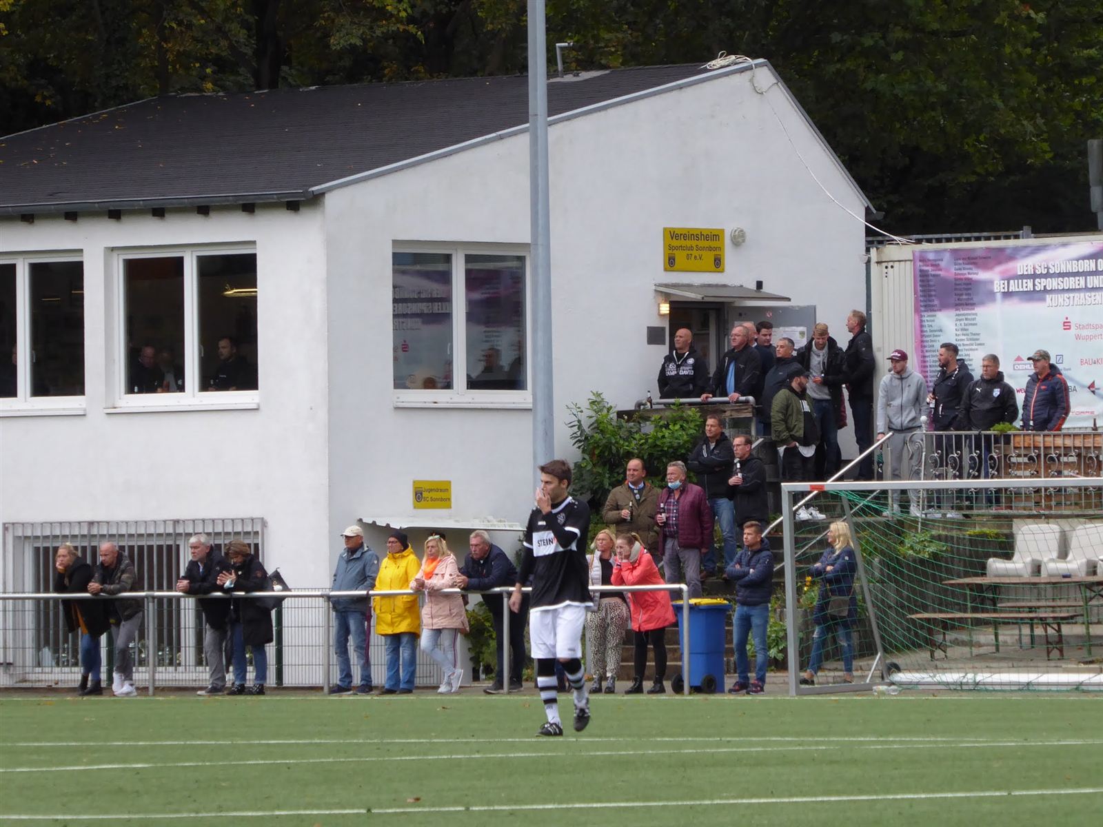 Vereinsheim mit Zuschauern am Spielfeldrand